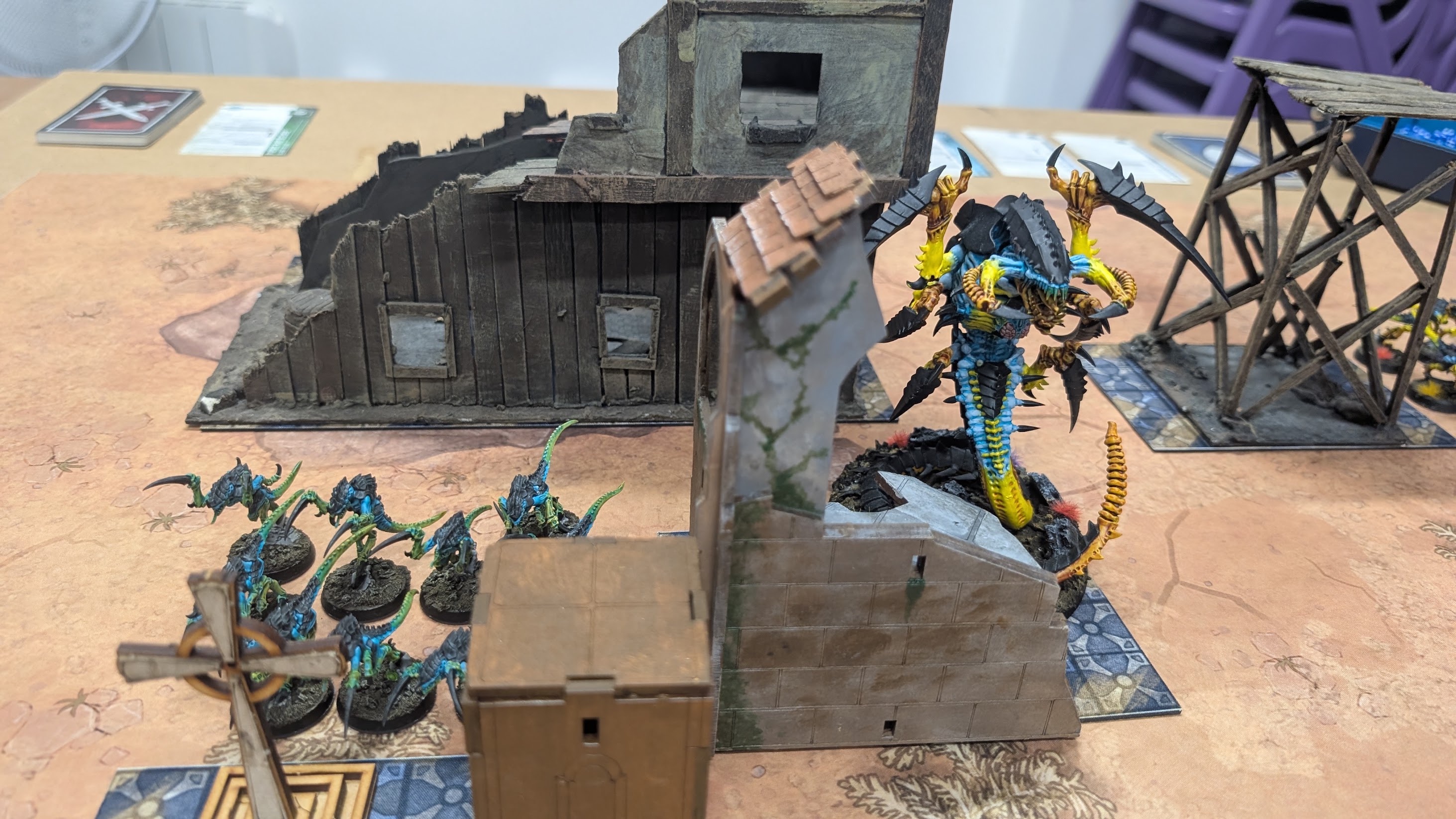 A yellow and teal winged Hive Tyrant rears up beside a ruined building