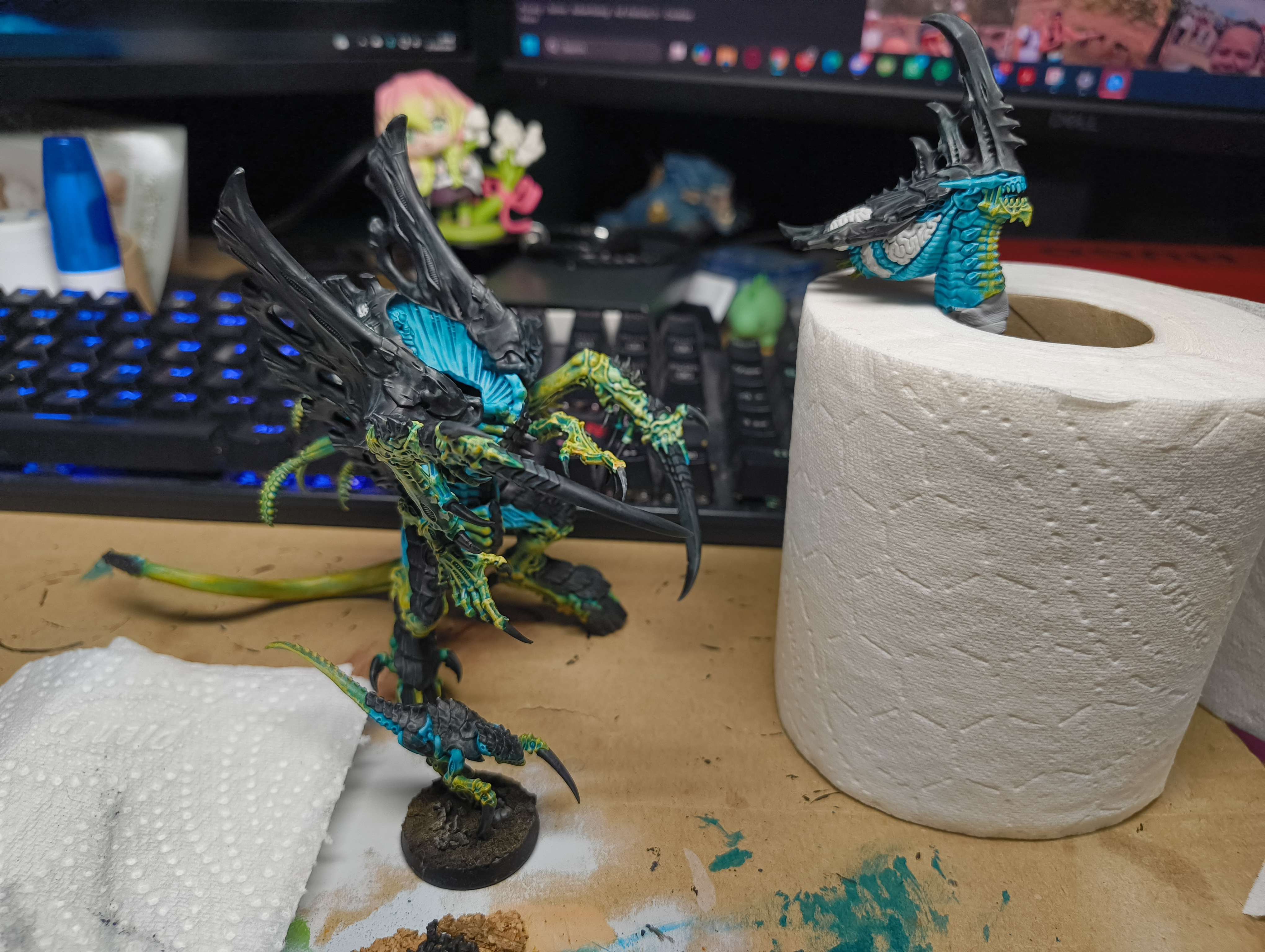 A teal and black winged Tyranid on a painting desk beside a second part-painted head